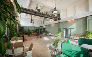 A general view of the large hall at Maják Zdiby with green walls and a concrete floor. In the foreground, there is a retro seating area with theater seats and houseplants. A tubular doublepower SEA light fixture is mounted on the right wall above the glass door.