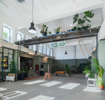 A view into the hall of Maják Zdiby, focusing on a central steel crane beam with a green backlit 'KINO' sign. A tubular doublepower SEA light fixture is mounted on the side wall below the beam, illuminating the area under the gallery. Wooden seating and plants are visible on the floor.