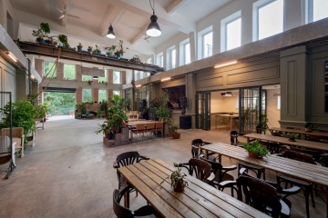 A view into the hall of Maják Zdiby with long, massive wooden tables and chairs for guests. Above them is a row of tall windows. The space is also pleasantly lit by tubular doublepower SEA light fixtures placed along the sides of the room.