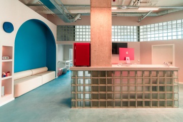 Modern reception area of the Mama Clinic in Prague with a distinctive design. In the foreground is a reception desk made of transparent glass blocks, behind which stands a massive concrete pillar, a pink iMac, and a red cabinet. On the left, there is a large, deep blue arched alcove with a built-in beige sofa. The entire space features a light blue floor and pastel pink walls. On the open industrial ceiling, slim linear LED luminaires by doublepower CELL are installed among the exposed ventilation ducts.