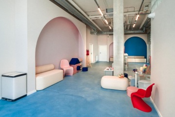 Bright and playful waiting room interior at Mama Clinic in Prague featuring a children's corner. The space is dominated by a turquoise-blue poured floor, raw concrete pillars, and pastel pink walls. In the foreground is a kids' play area with toys and a designer bright red chair. In the background, another blue decorative arch is visible on the wall alongside comfortable seating for adults. Lines of minimalist doublepower CELL luminaires are suspended from the exposed concrete ceiling, illuminating the space evenly.
