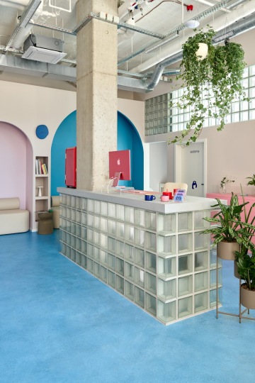 Side view of the glass block reception desk at Mama Clinic Prague. A pink monitor is placed on the counter, with pastel pink walls featuring blue accents and a white door visible in the background. The interior is enlivened by green houseplants in pots on the floor and hanging from the ceiling. Linear doublepower CELL luminaires are attached to the open industrial ceiling with an exposed air conditioning unit and piping, completing the modern look of the clinic.